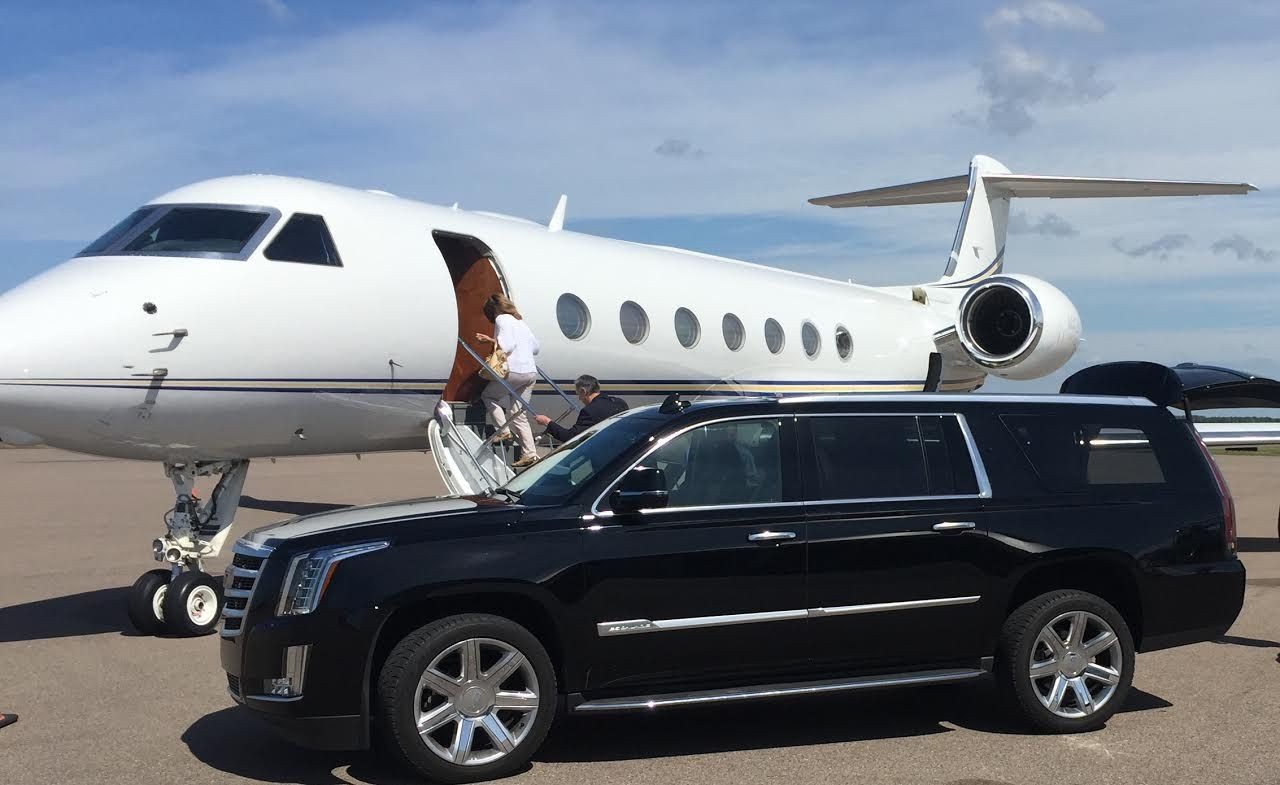 White private jet parked on tarmac with people boarding, next to a black luxury SUV.