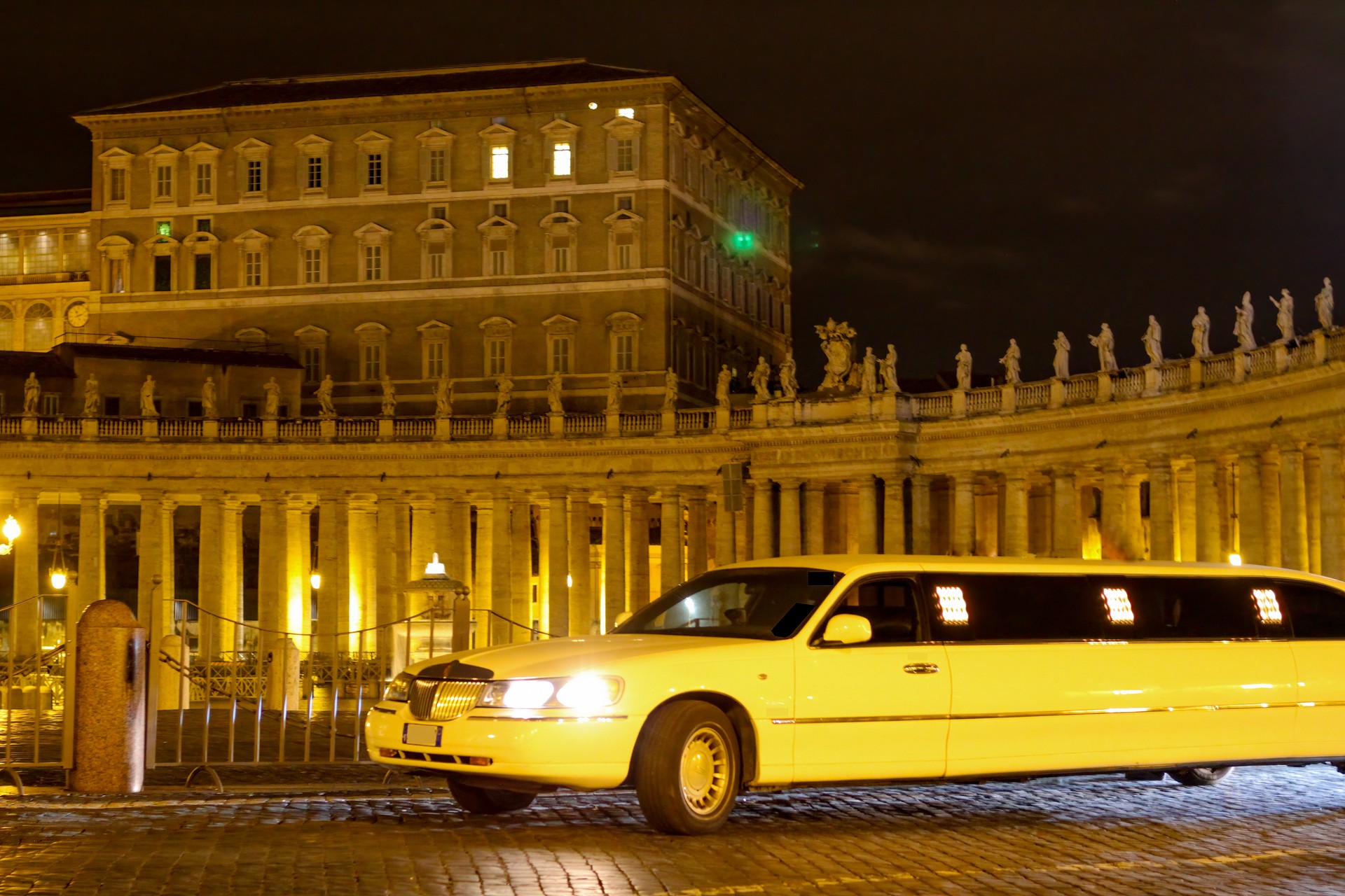 A white stretched limo is parked on a square at night with its headlights on